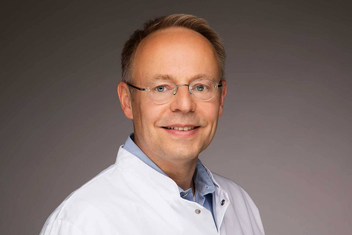 Ein lächelnder Mann mit kurzen hellbraunen Haaren und Brille, der einen weißen Laborkittel und ein hellblaues Hemd trägt, posiert vor einem schlichten Hintergrund mit Farbverlauf.